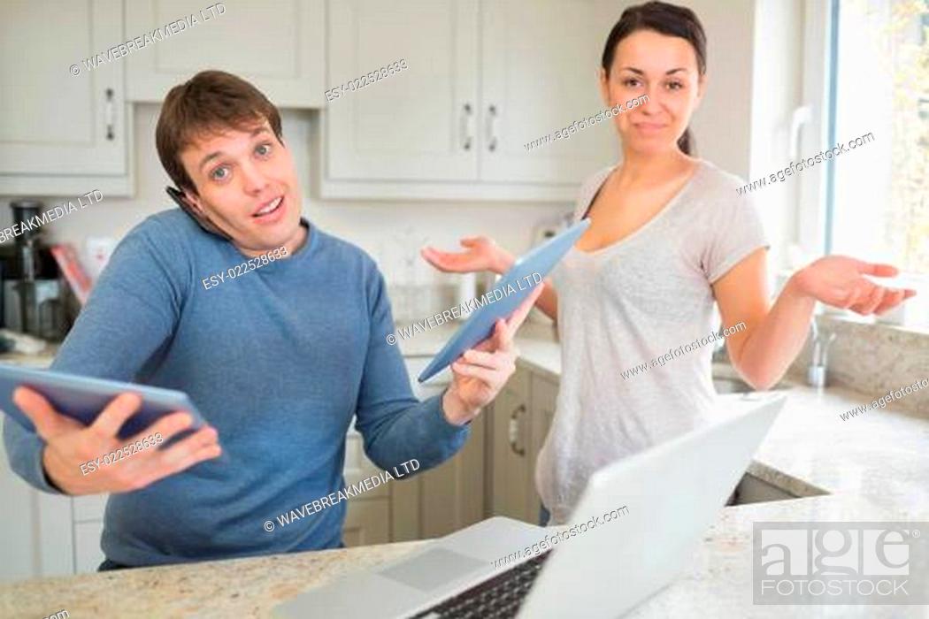 Stock Photo: Busy man using two tablets and laptop with wife holding hands up questioning in kitchen.