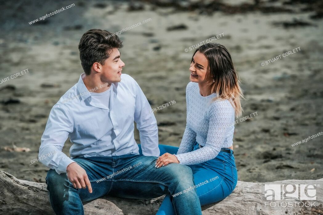 Stock Photo: Portrait of a young couple sitting on a log at the beach; Wellington, North Island, New Zealand.