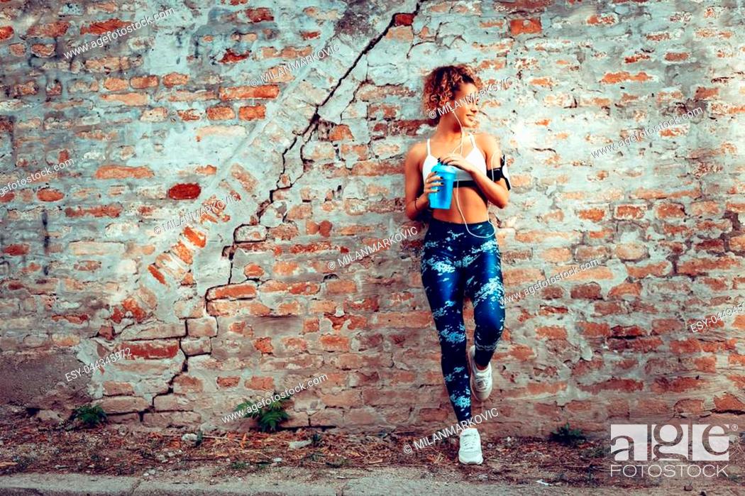 Stock Photo: Young muscular woman with headphones standing up against the wall and resting after jogging.