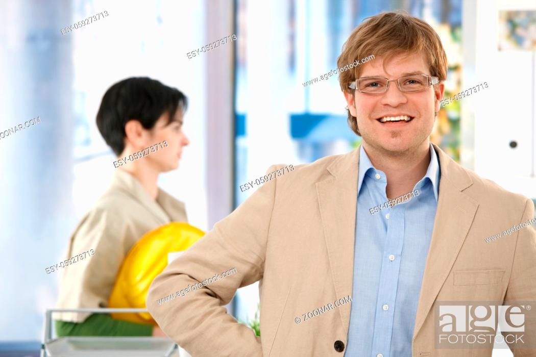 Imagen: Portrait of happy young architect standing in office, colleague walking in background.