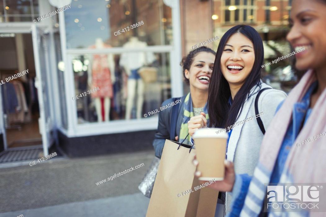 Stock Photo: Laughing women friends walking along storefront with coffee and shopping bags.