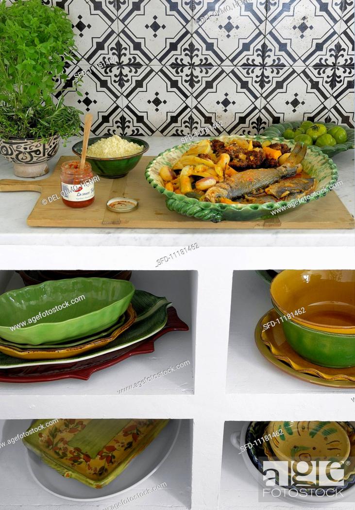 Stock Photo: A kitchen shelf unit holding stacked ceramic crockery; on the top, a plate of oven-baked fish and vegetables and a jar of harissa sauce.