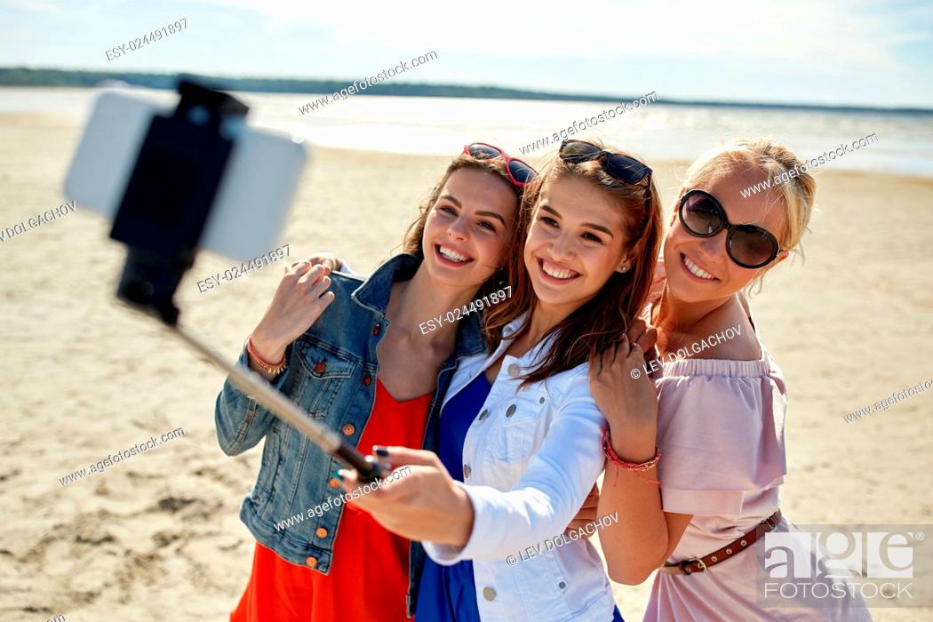 Stock Photo: summer vacation, holidays, travel, technology and people concept- group of smiling young women taking picture with smartphone on selfie stick on beach.