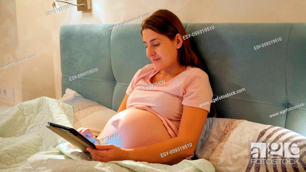 Stock Photo: Smiling pregnant woman wearing pajamas working on tablet and browsing internet before going to sleep in bed at night.