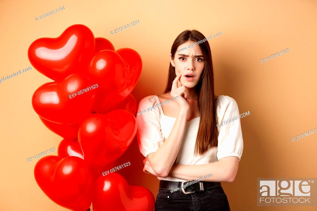 Stock Photo: Valentines day and love concept. Confused and shocked girl staring displeased at camera, standing near romantic red hearts balloons on beige background.