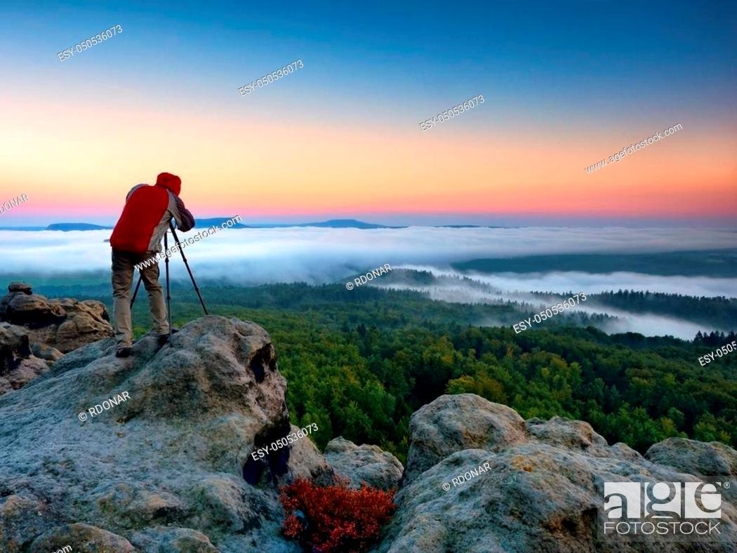Imagen: Photographer takes photos with mirror camera and tripod on peak of rock. Fall fogy landscape, spring orange pink misty sunrise in beautiful valley below.