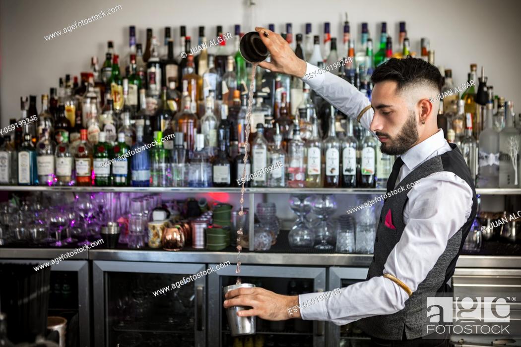 Imagen: Side view of young bearded male barkeeper in elegant uniform pouring alcohol drink from shaker into stainless cup while preparing order at bar counter.
