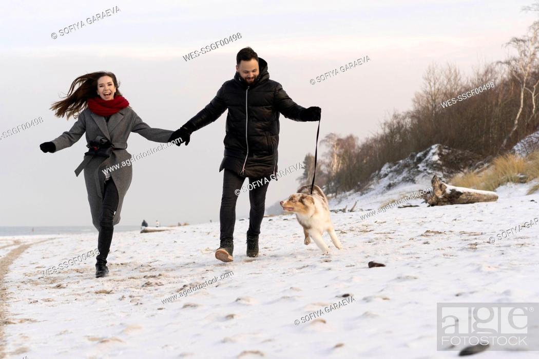 Imagen: Happy young couple running with dog on snow at sunset.