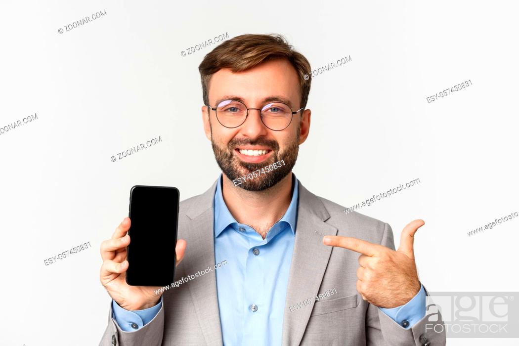 Stock Photo: Close-up of confident businessman in glasses and gray suit, pointing finger at mobile phone screen and smiling proud, standing over white background.