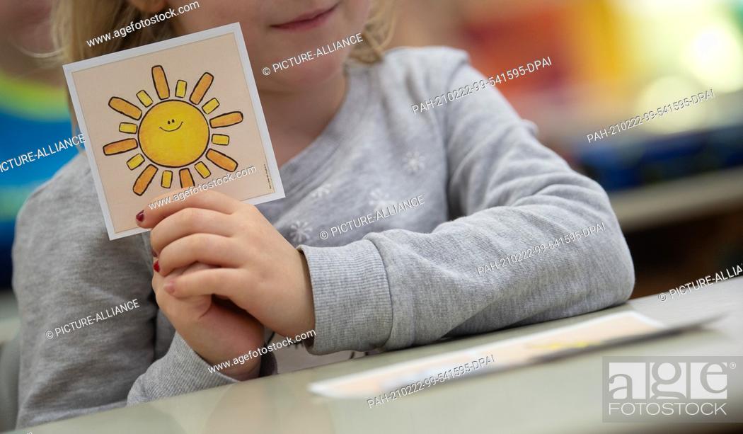 Stock Photo: 22 February 2021, Baden-Wuerttemberg, Hemmingen: A girl in a first grade class at an elementary school with rotating classes is holding a card with a sun on it.