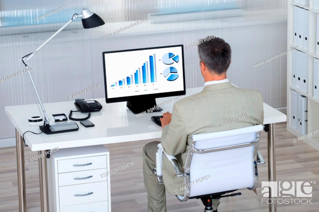 Imagen: Rear view of businessman analyzing financial graphs on computer at desk in office.