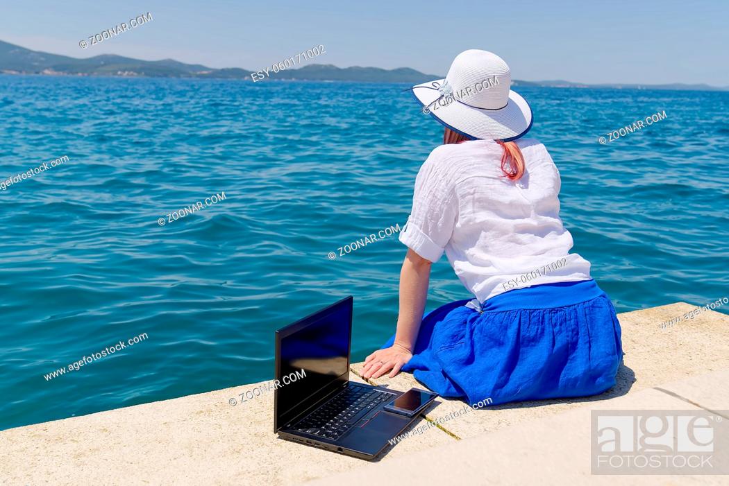 Stock Photo: Work from anywhere. Side view of young woman, female freelancer in straw hat working on laptop while sitting on the beach.