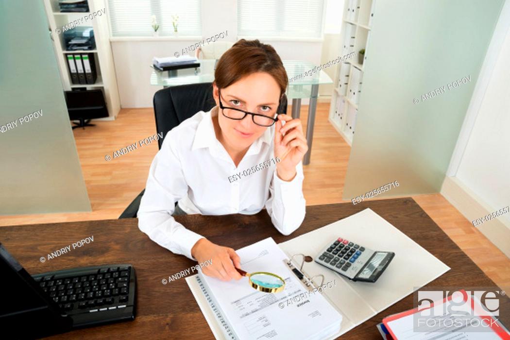 Stock Photo: Young Businesswoman With Invoice And Magnifying Glass At Desk In Office.
