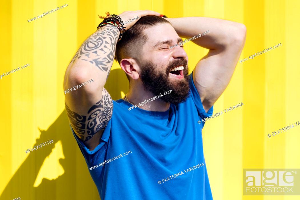 Stock Photo: Laughing young man with tattoo standing in front of yellow wall.
