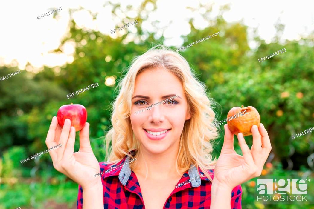 Stock Photo: Beautiful young blond woman in checked red shirt harvesting apples, holding in her hands one rotten and one good apple.