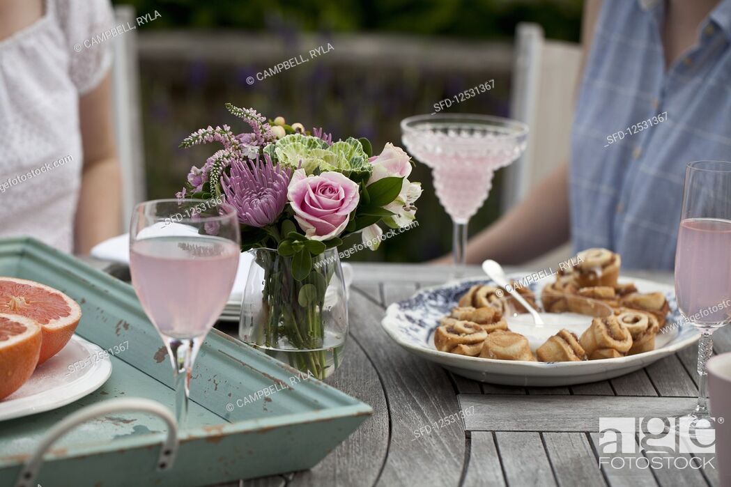 Stock Photo: Two women sitting at an outdoor brunch table with flowers, grapefruit halves, glasses of sparkling strawberry lemonade, cinnamon buns and icing.