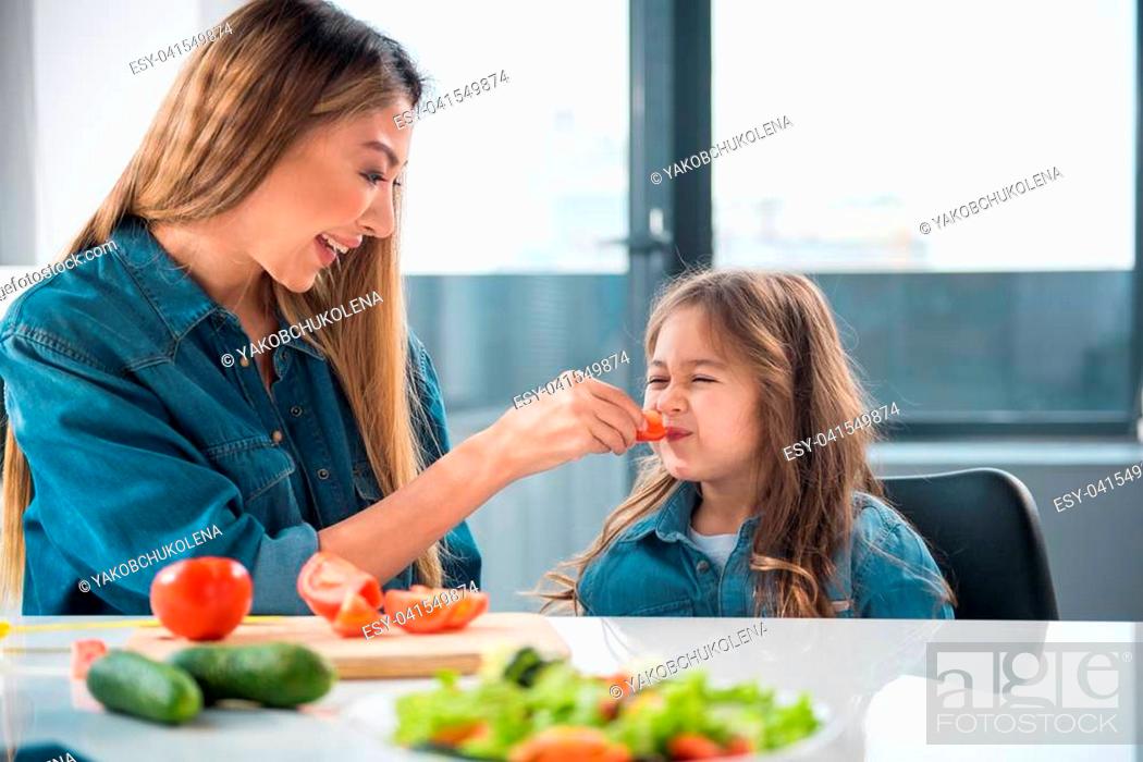 Stock Photo: Smiling woman is giving piece of fresh vegetable to her daughter. The girl closing her mouth with aversion.