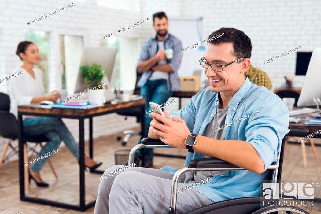 Photo de stock: Disabled person in the wheelchair works in the office. He uses his smartphone and smiling. His colleagues work nearby.