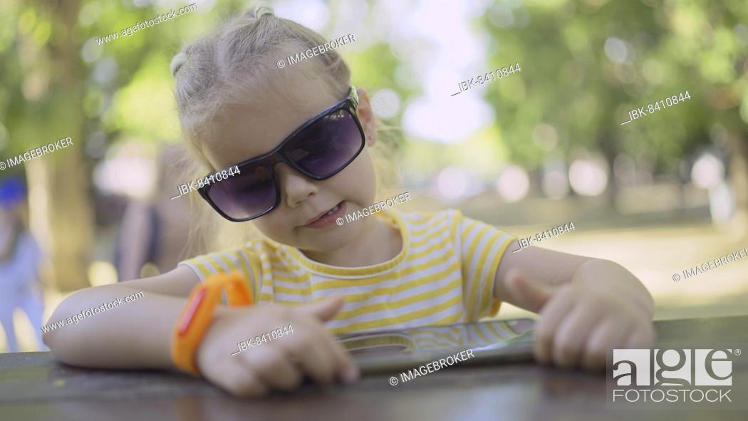 Stock Photo: Cute little girl in moms sunglasses listens to music on a mobile phone and sings along. Close-up portrait of child girl sitting in sunglasses on city park and.