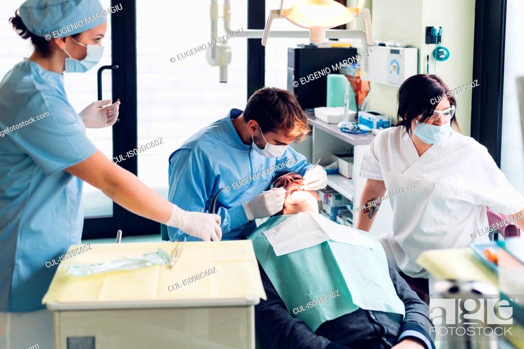 Imagen: Dentist looking into male patient's mouth, dental nurses preparing equipment.