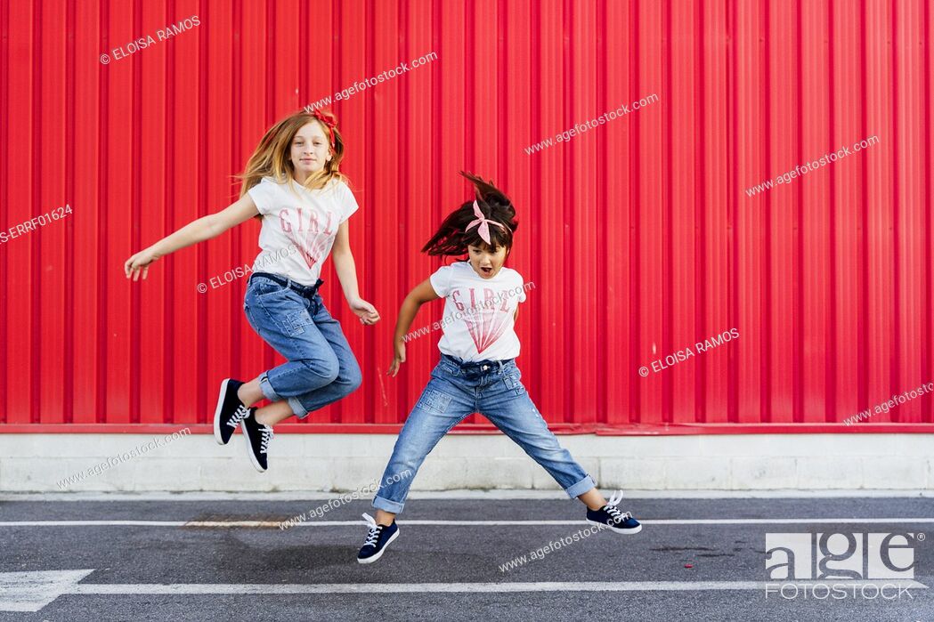 Stock Photo: Sisters jumping on street in front of red wall.