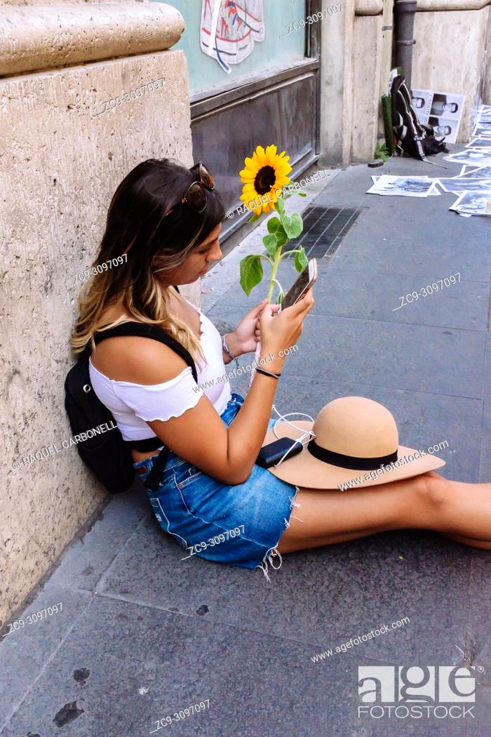 Stock Photo: Young girl sit on the street looking at her mobile phone while holding an small sunflower in her hand, Rome, Lazio region, Italy.