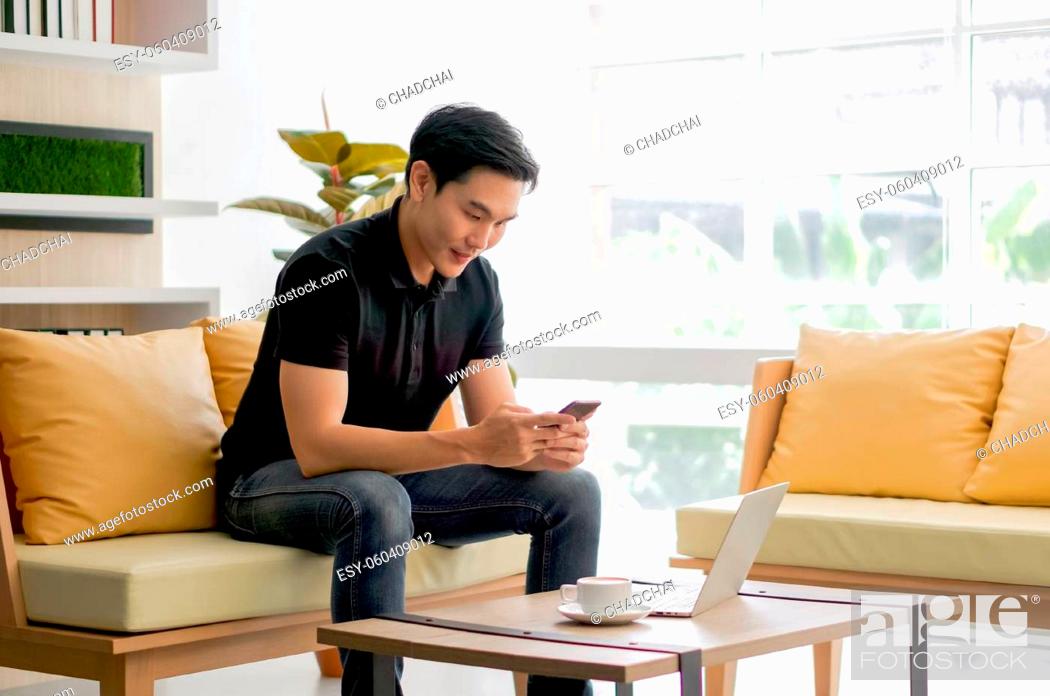 Stock Photo: Young asian man reading text message on mobile phone while working with laptop computer in the living room. A cup of coffee was placed on the table next to him.