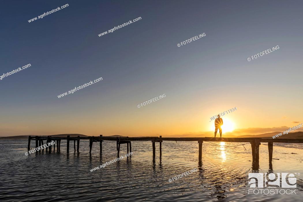 Imagen: Silhouette of woman standing alone on coastal jetty at sunrise.