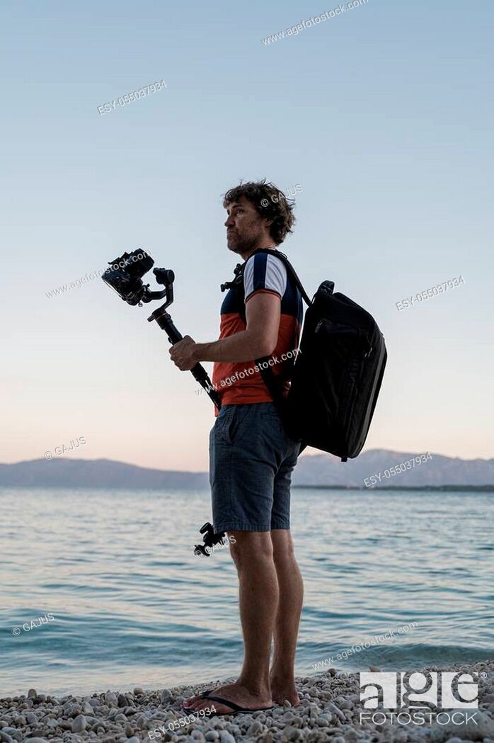Stock Photo: Young male photographer standing on pebble beach in the evening, holding his dslr camera on gimbal stick looking for a perfect spot to shoot a video.