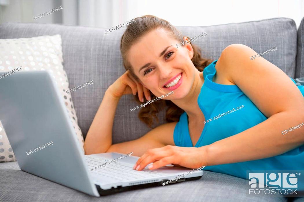 Stock Photo: Smiling young woman laying on sofa with laptop.