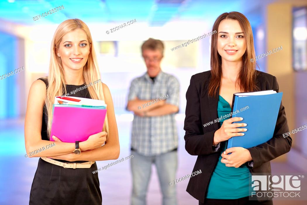Stock Photo: Two girls in the office with folders with papers in hand. Young women are chatting and laughing. Their angry boss watches from behind.