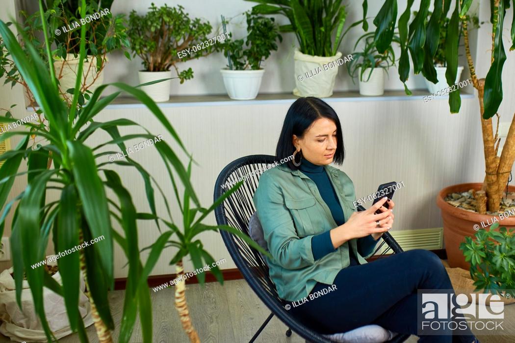 Stock Photo: Woman chatting using smartphone, sitting on armchair at home, types message with happy smile, Concept of home garden, Biophilia design.