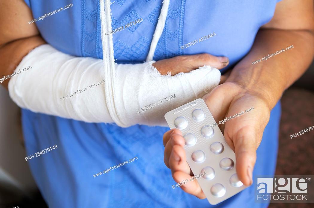 Stock Photo: A senior retired woman with a rewound arm in a cast and a bandage holds anesthetic pills. Blow, fracture, bones, hospital || Model approval available.