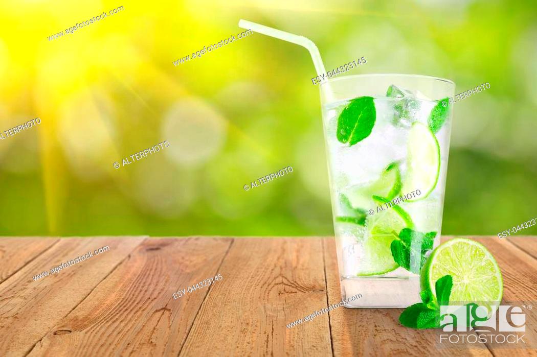 Stock Photo: glass of cold lemonade with lemon slices, mint leaves and ice cubes on wooden table against green blurred natural background.
