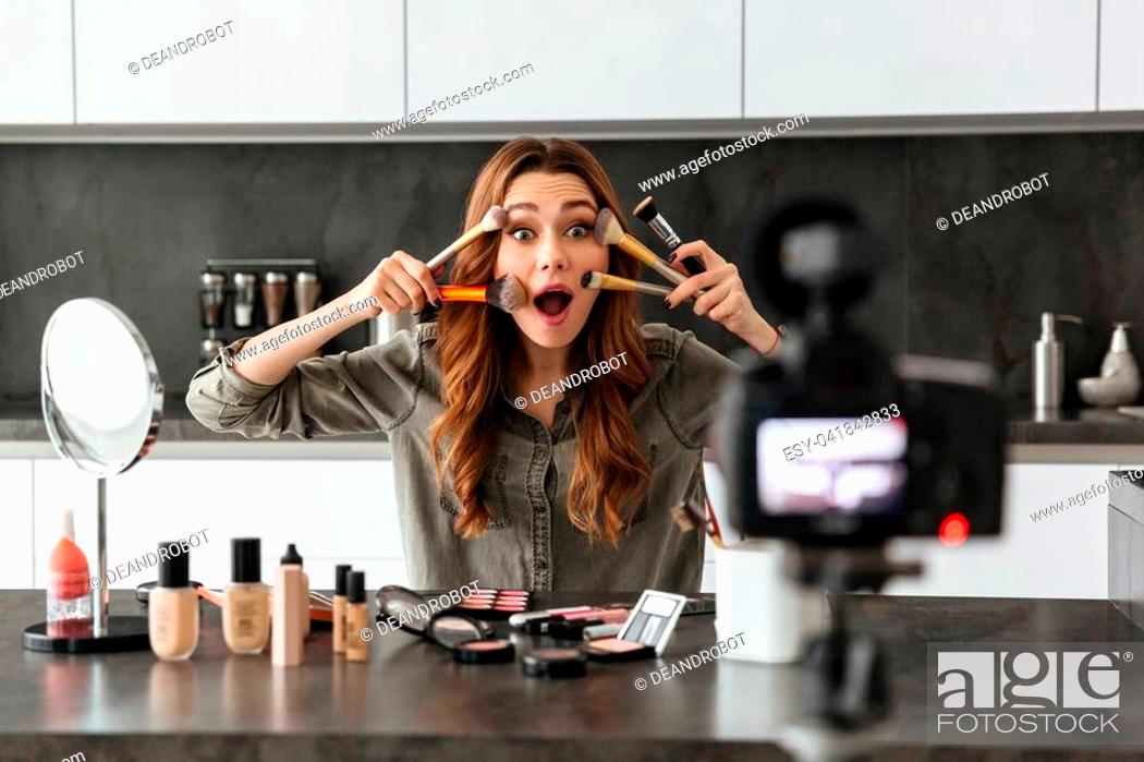 Stock Photo: Happy young girl recording her video blog episode about new cosmetic products while sitting at the kitchen table at home and applying make-up.