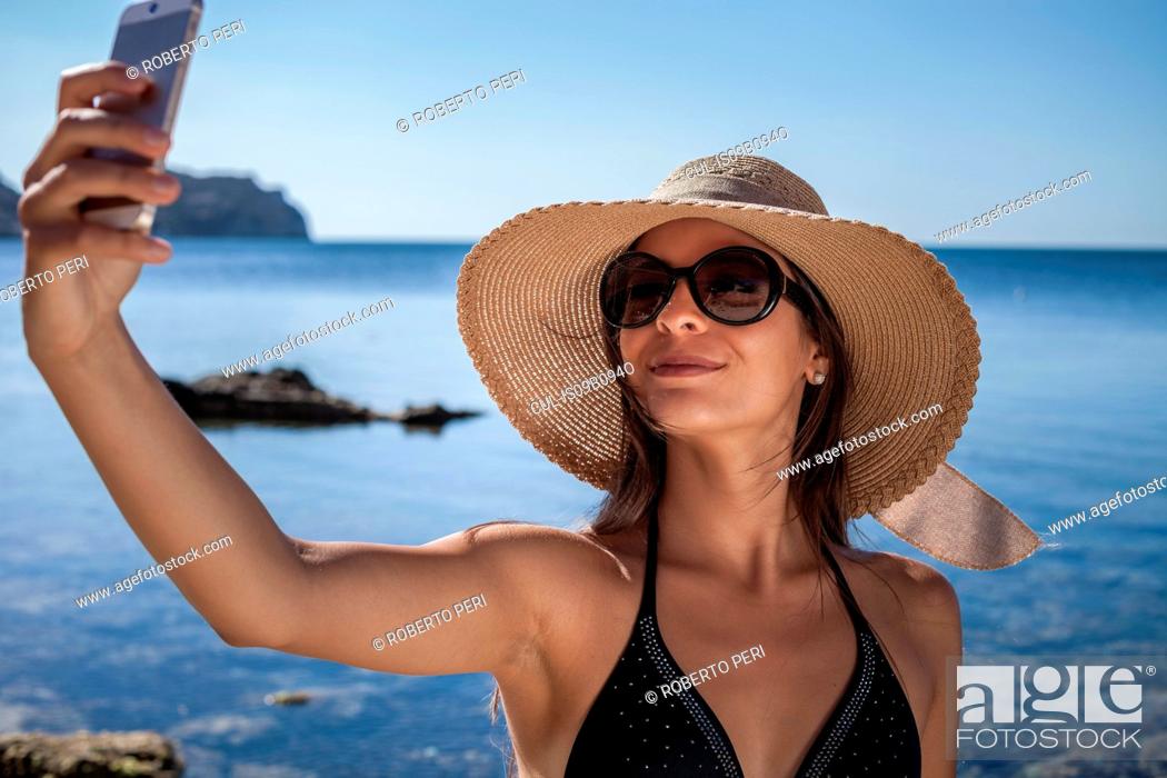 Stock Photo: Young woman wearing sunhat on beach taking smartphone selfie, Villasimius, Sardinia, Italy.