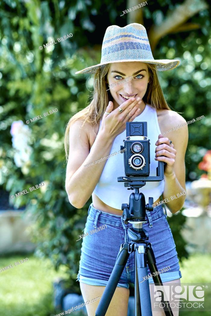 Stock Photo: Portrait of a young beautiful caucasian woman in her 20's with a gesture of shyness wearing a hat and photographing with an old vintage camera on a tripod.