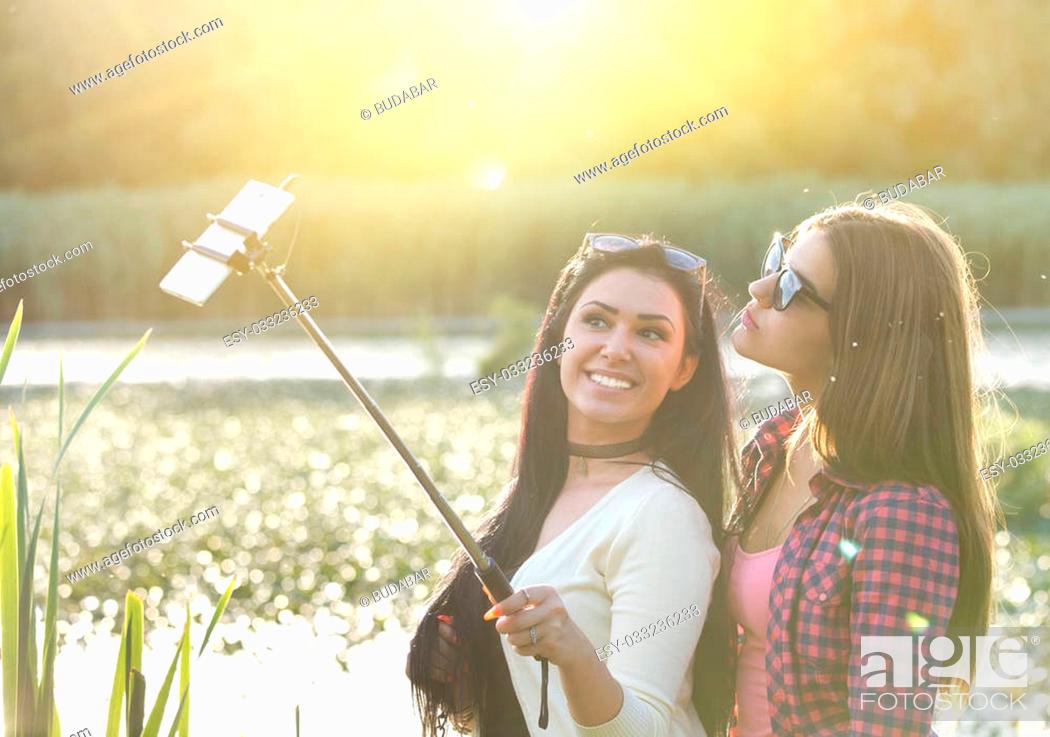 Stock Photo: Two pretty teenage girls taking selfie on mobile phone with stick in the nature beside river. Positive emotions.