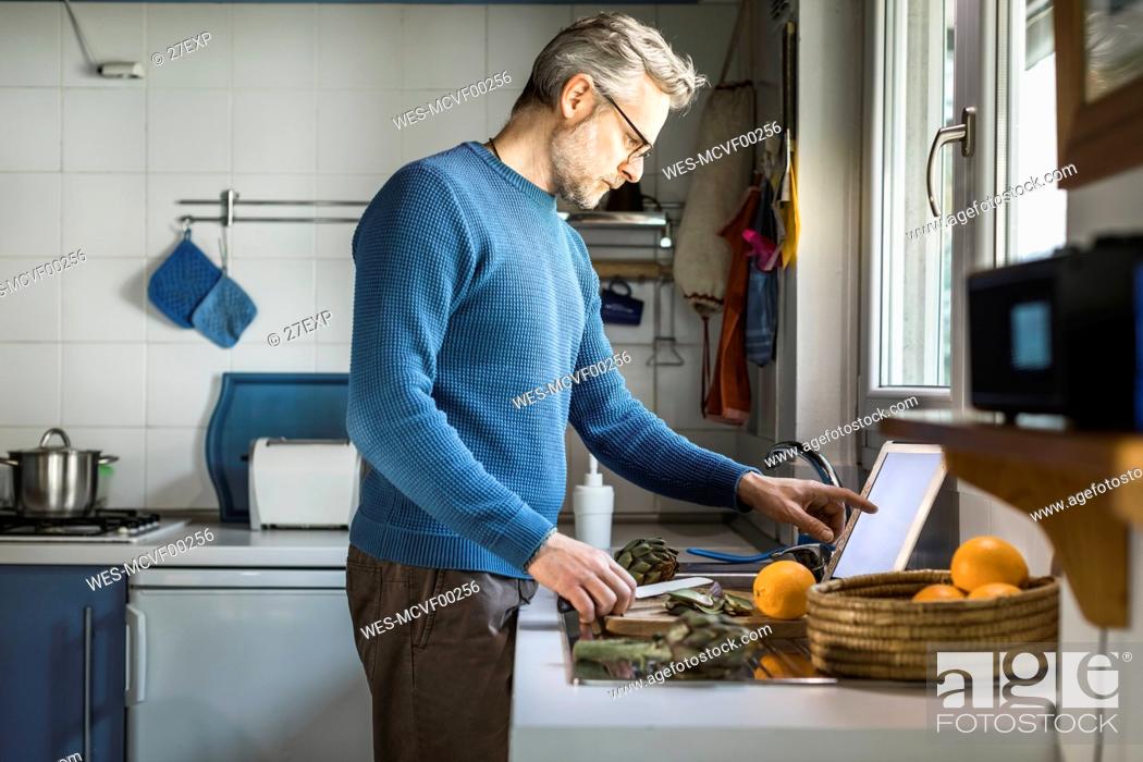 Stock Photo: Mature man preparing artichokes in his kitchen using digital tablet.