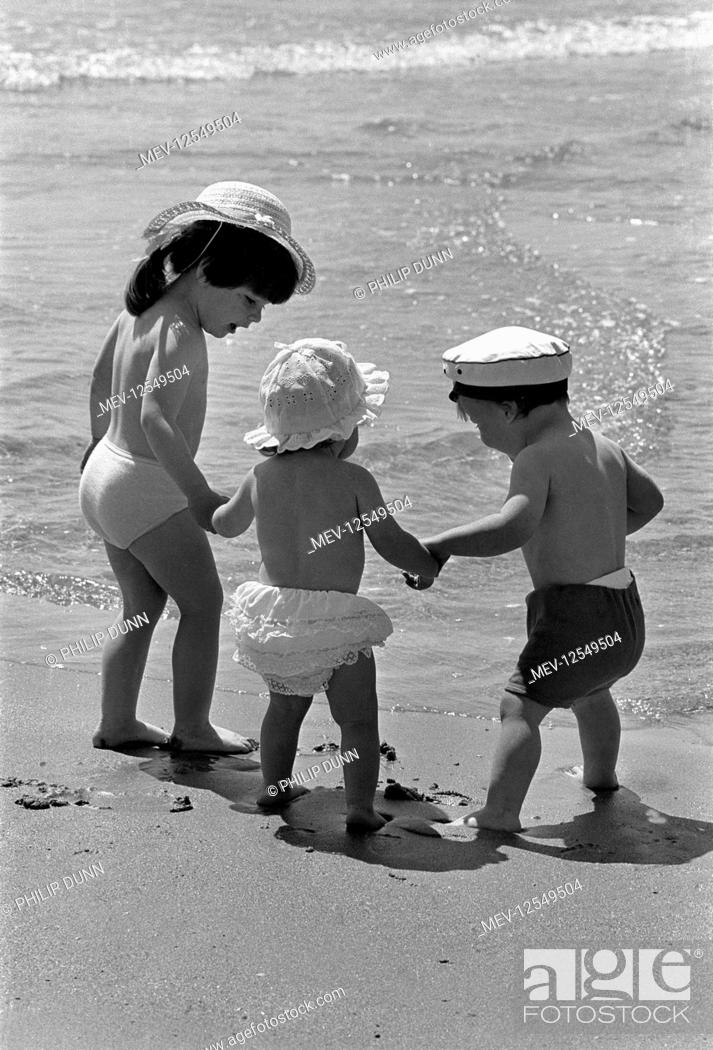 Stock Photo: Three small children wearing sun hats hold hands in the sunshine and walk to the sea on the beach at Tenby, Pembrokeshire, Wales.