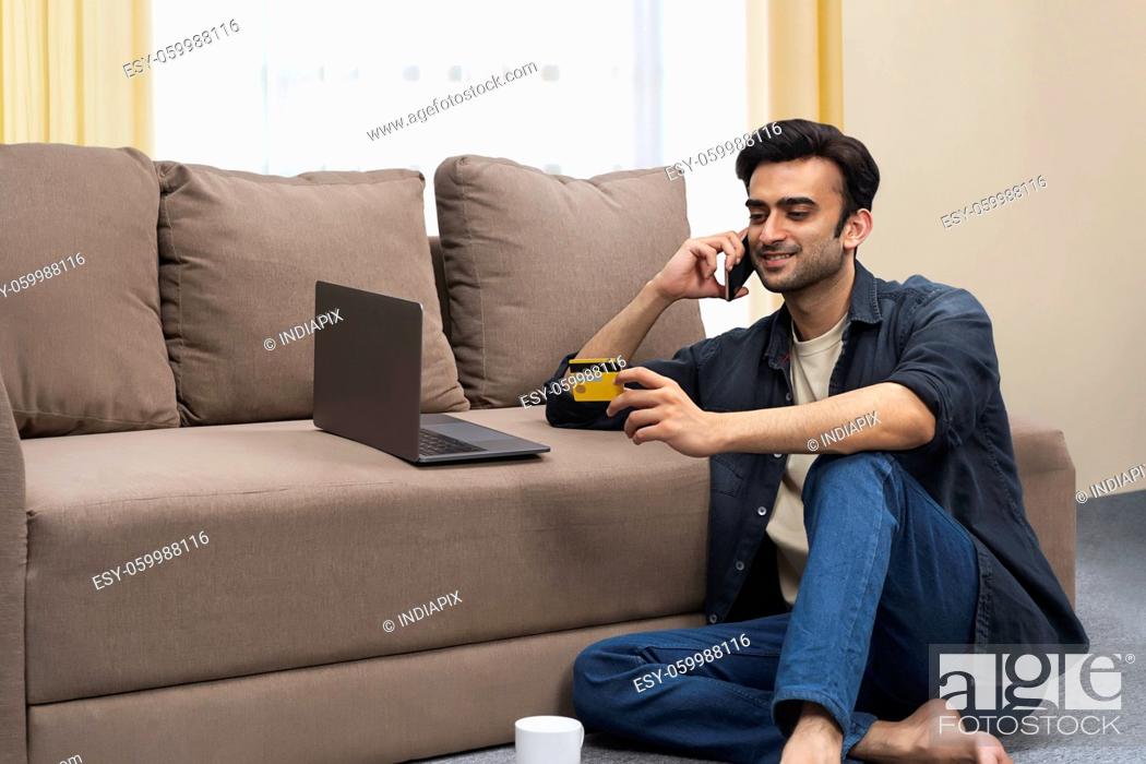 Stock Photo: A young man with laptop holding credit card talking on mobile in his living room.