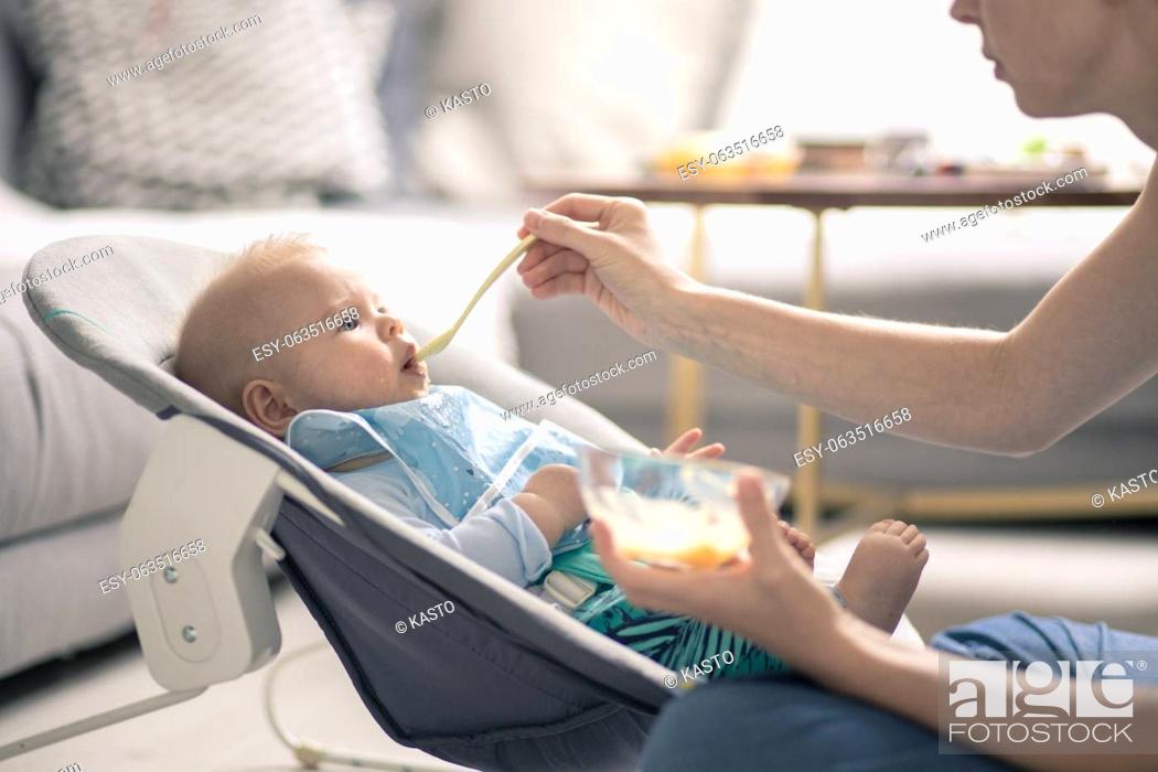 Stock Photo: Mother spoon feeding her baby boy child in baby chair with fruit puree. Baby solid food introduction concept.