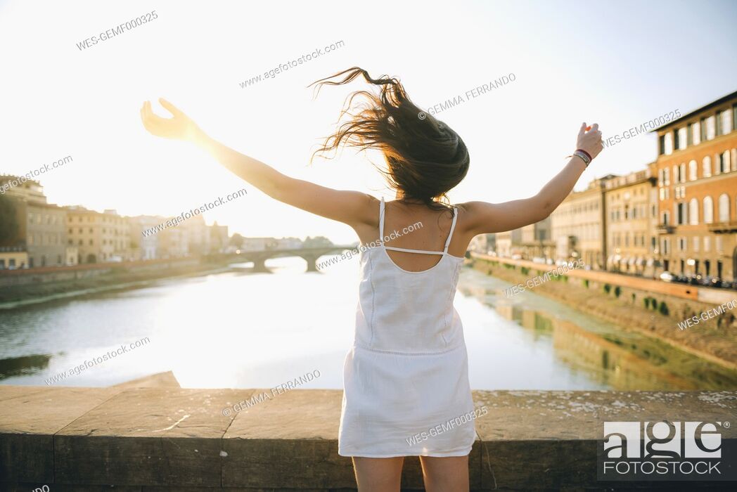 Imagen: Italy, Florence, back view of happy woman wearing white summer dress standing on a bridge at sunset.