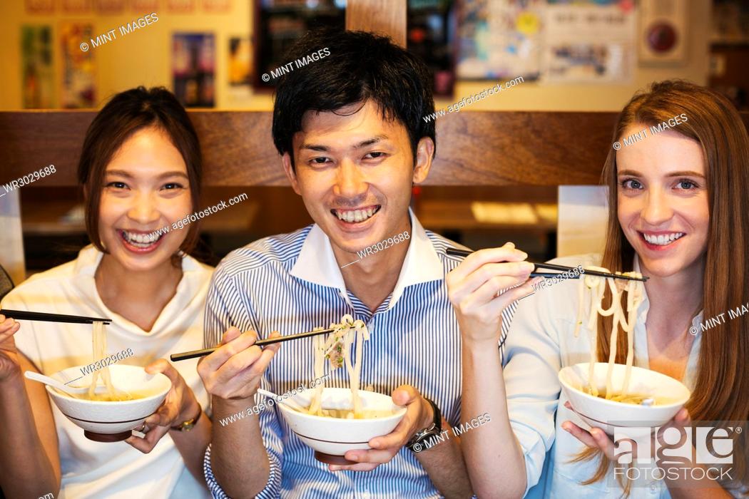 Stock Photo: Three smiling people sitting sidy by side at a table in a restaurant, eating from bowls using chopsticks.
