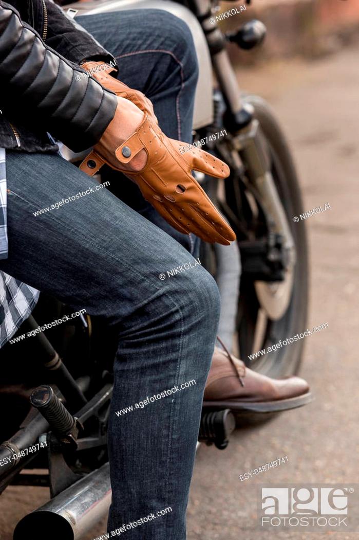 Stock Photo: Close-up of a handsome rider biker man hand puts on a glove while sit on classic style cafe racer motorcycle. Bike custom made in vintage garage.