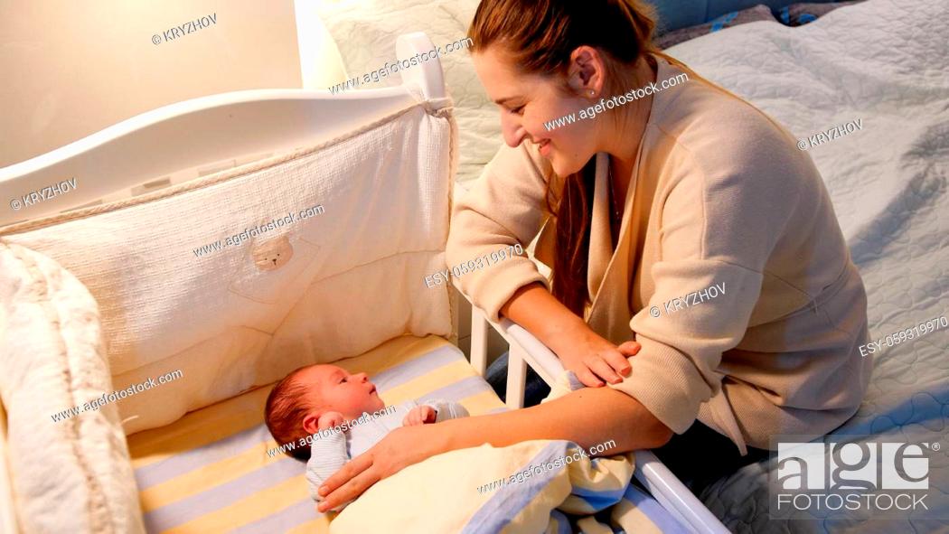 Stock Photo: Happy smiling mother rocking crib and looking on her newborn baby boy lying in bed at night. Concept of happy parenting and family happiness.