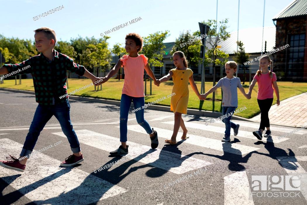 Stock Photo: Front view of a diverse group of elementary school pupils crossing an empty road together, half way across a pedestrian crossing, holding hands on a sunny day.