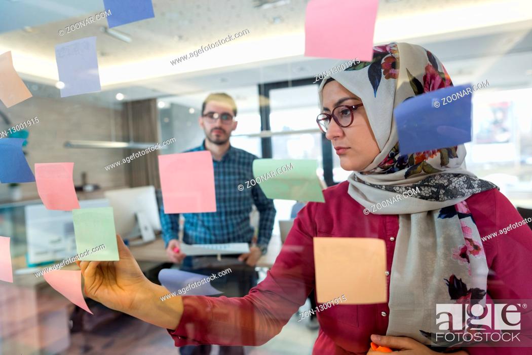 Stock Photo: Beautiful Arab woman wearing hijab putting sticky notes on glass in modern office.