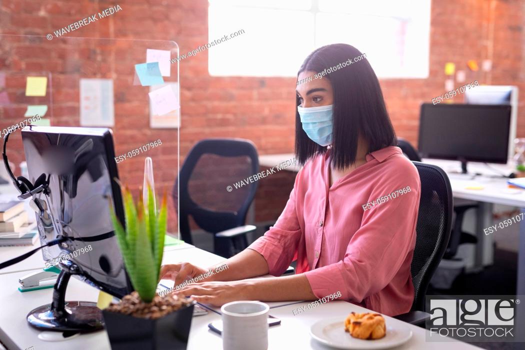 Stock Photo: Mixed race businesswoman sitting in office in front of computer wearing face mask.