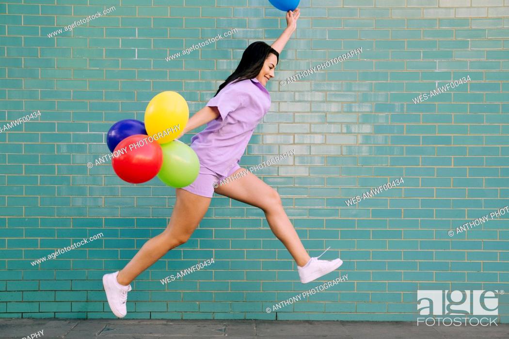 Stock Photo: Carefree young woman jumping with balloons by wall.